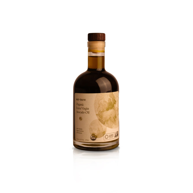 A clear glass bottle of west~bourne Organic Extra Virgin Avocado Oil with a wooden cap and beige label sits on a white background—ideal for high-heat cooking and an essential for any sustainable kitchen.