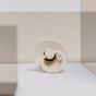 A beige and brown baby pacifier stands upright on a white surface, with a soft-focus background in neutral tones and geometric shapes.