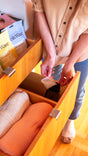 A person in a beige shirt organizes a wooden drawer, placing a small item inside alongside folded orange and beige clothes. Surrounding them are Fresh Wave Odor Removing Packs for Small Spaces, made with plant-based ingredients. The floor is wooden, and they wear cozy socks.