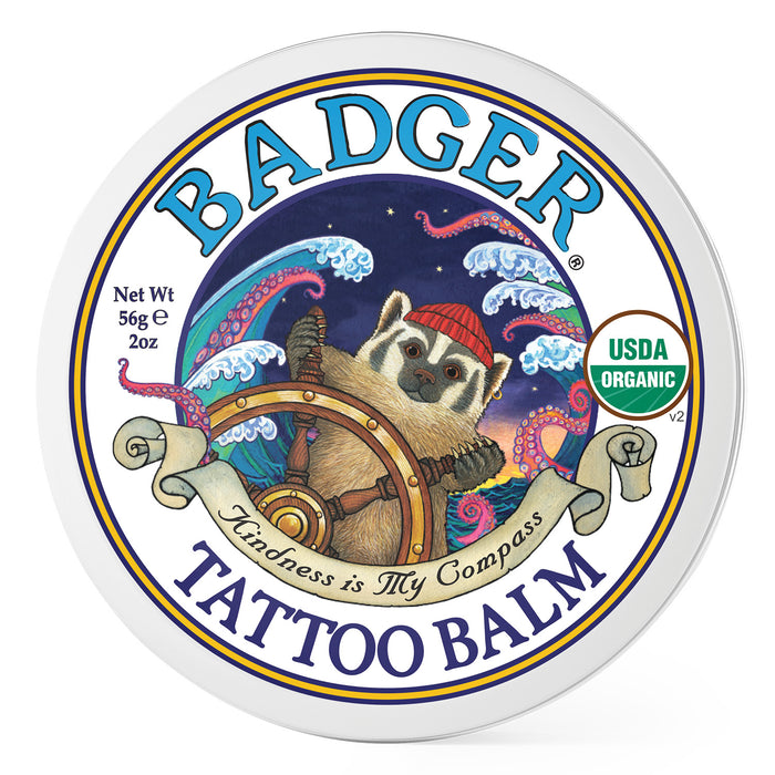 A round tin of Badger Tattoo Balm features an illustrated badger wearing a red knit hat, steering a ships wheel with colorful waves behind it. Text reads Kindness is My Compass and shows USDA Organic seal.