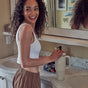 A smiling woman with curly hair and a white tank top stands by a bathroom sink, holding a dropper above a large, light-colored cup on the counter.