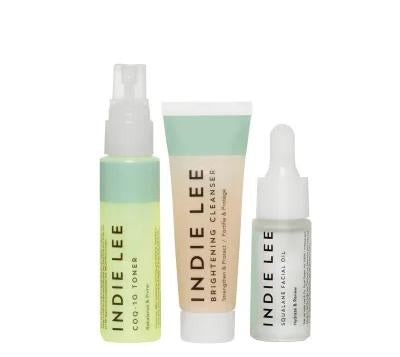 Three Indie Lee skincare products: a green-capped spray toner, a tube of brightening cleanser, and a small dropper bottle of squalane facial oil, all with minimalist packaging.
