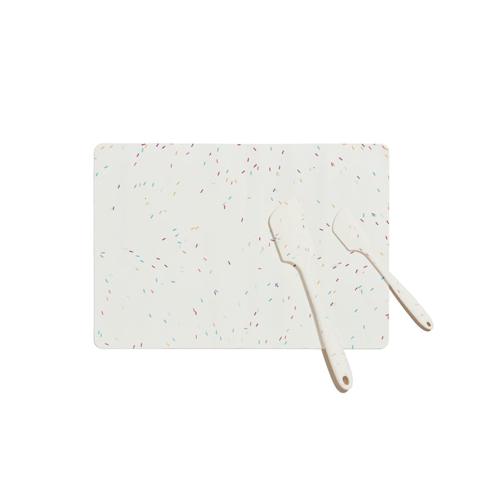 A white rectangular cutting board with colorful confetti-like specks, accompanied by two white spatulas, all set against a plain white background.