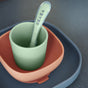 A green cup with a matching spoon inside sits in an orange bowl, which is placed on a blue plate. The word BEABA is visible on the spoon. The items appear to be made of silicone.
