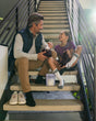 A man and a girl sit on indoor stairs, smiling and putting deodorizers into their shoes. Both appear relaxed and happy, with sunlight streaming in from the side. A Fresh Wave odor-removing packet sits beside them.