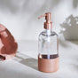 A clear glass soap dispenser with a rose gold pump and a blush pink base sits on a light marble surface. Sunlight casts soft shadows, and a pink cloth is partially visible on the left side.