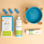 Flat lay of Oxyfresh pet dental care products, including a dental kit box, water additive bottle, toothpaste tube, two finger toothbrushes, and a blue pet bowl on a beige background.