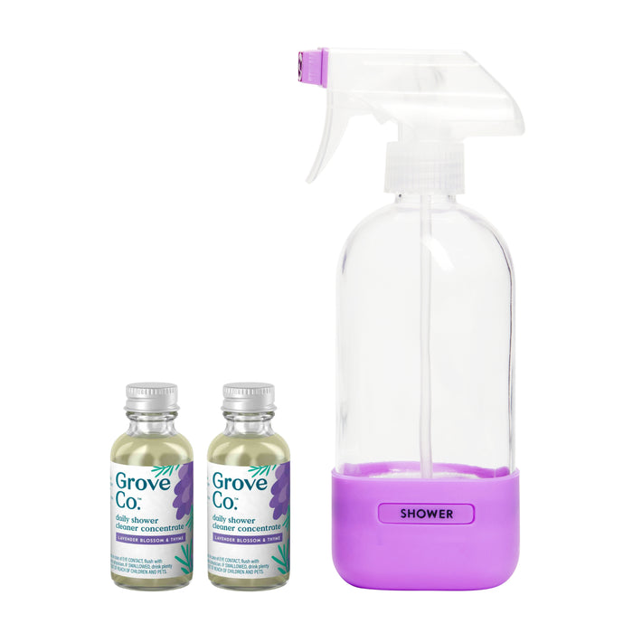 Two small bottles of Grove Collaborative Daily Shower Cleaner Concentrate sit beside a clear spray bottle with a purple base labeled Shower. The cleaner bottles feature green and lavender labels adorned with purple leaf designs.