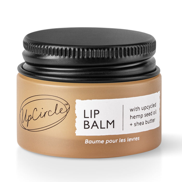 A small brown jar with a black lid labeled “UpCircle Lip Balm” containing upcycled hemp seed oil and shea butter. The label is white with black text, and there is French text at the bottom.
