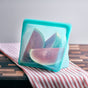 A reusable silicone bag containing watermelon slices sits on a red-and-white striped cloth on a wooden surface with a light gray background.