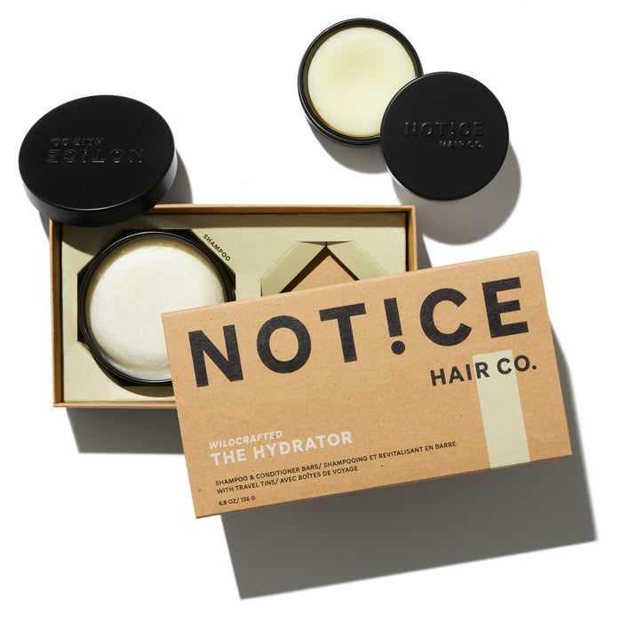 A box labeled NOT!CE HAIR CO. THE HYDRATOR with a shampoo and conditioner bar set is open, showing two round bars, one in a tin. Another tin with a round bar is nearby, all on a white background.