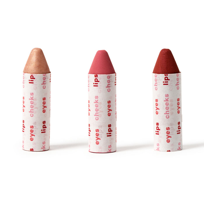 Three crayon-shaped makeup sticks stand upright in a row. Each has different shades—beige, pink, and red—and is wrapped in white paper printed with the words lips, cheeks, and eyes in red text.