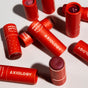 Several red tubes of Axiology multi-stick color cream are scattered on a white surface, some with lids off revealing the product inside. The tubes have white branding and heart patterns.