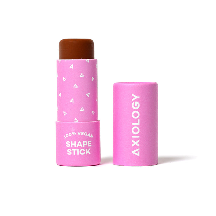 A brown cosmetic shape stick in a pink tube with white triangle patterns, labeled 100% Vegan Shape Stick. The cap, which reads Axiology, is standing next to the open stick.
