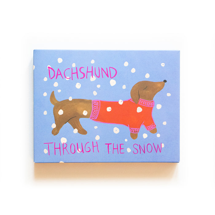 A drawing of a dachshund wearing a red sweater, surrounded by falling snow on a blue background. Pink text reads Dachshund Through The Snow.