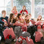 A group of kids in pajamas sit on a couch, smiling and holding large gift bags with festive patterns. Sunlight streams through the windows behind them, and they appear to be celebrating together.