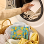 A person in a beige sweater places a white sheet into a washing machine filled with white towels. In front, a wicker basket holds folded towels and Grove Co.s Laundry Sheets - Spring Collection, designed to combat plastic pollution.