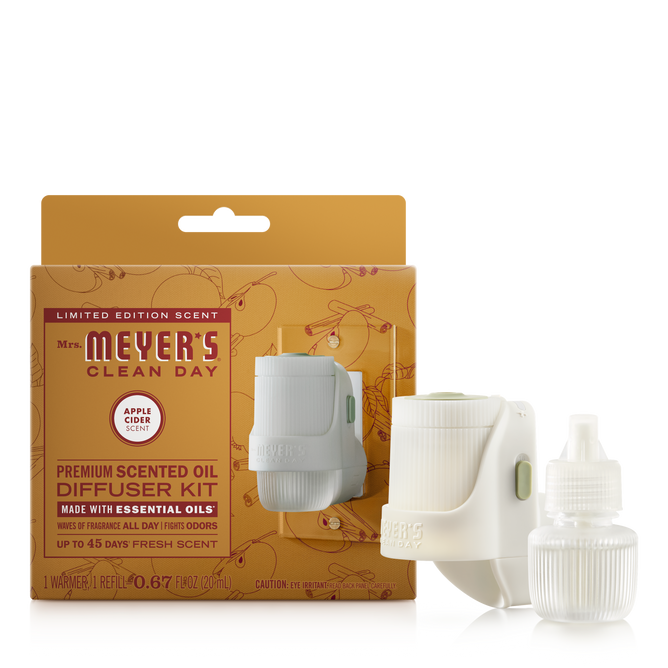 Mrs. Meyer's Premium Scented Oil Diffuser Kit includes a white plug-in diffuser, a clear scented oil refill, and comes in a brown and orange box featuring apple illustrations.