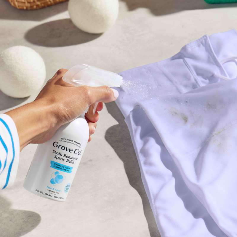 A hand sprays Stain Remover Spray by Grove Co., filled with natural cleaning enzymes, onto a stained white cloth. On a light surface, wool dryer balls in sight, the lit scene reflects an eco-friendly cleaning ritual with this cruelty-free product.
