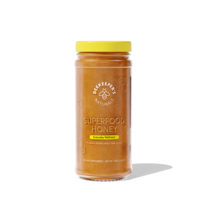 A clear jar with a yellow lid containing Beekeeper’s Naturals Superfood Honey. The label highlights Everyday Wellness with ingredients propolis, royal jelly, and bee pollen. The jar casts a faint shadow.