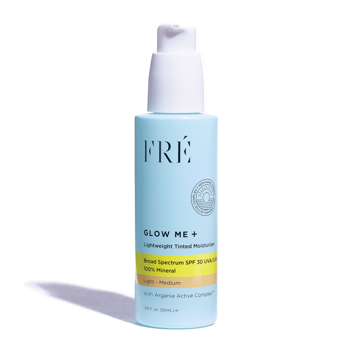 A light blue bottle of FRÉ Glow Me+ Lightweight Tinted Moisturizer with a pump dispenser, featuring broad spectrum SPF 30 and labeled as 100% mineral, light/medium shade, and containing argania active complex.