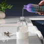 A hand pours liquid from a purple Grove Co. bottle into a clear dispenser bottle with a white base, set on a counter near a sink with a soap pump and a plant in the background.