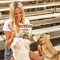A smiling woman sits on bleachers holding a bag of dog treats and a biscuit, while two dogs eagerly look at her, waiting for a treat.