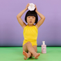 A young child in a yellow swimsuit sits on a green floor against a purple background, holding a white container above their head. A lotion bottle with a simple graphic design is placed beside them.