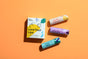Close-up of Plant Therapys Love Your Lips Trio Set on an orange background. Includes a yellow Love Your Lips box and three tubes: Vanilla Citrus (orange/pink), Lavender Lemonade (purple), Cool Mint (blue/turquoise); made with natural ingredients and featuring matching graphic designs.