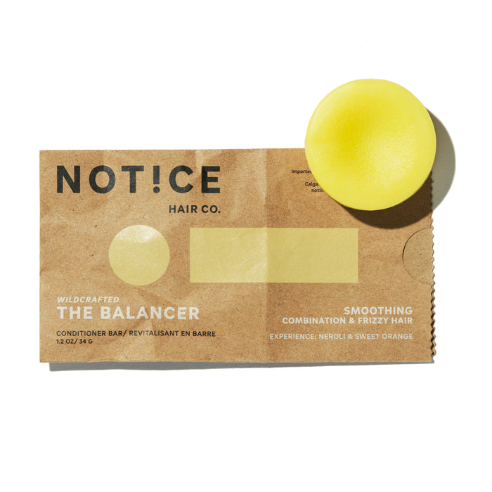 A yellow conditioner bar sits on top of a brown paper package labeled NOTICE Hair Co. The Balancer Smoothing Conditioner Bar, designed for combination and frizzy hair with neroli and sweet orange scent.
