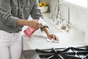 A person in a gray shirt and white pants sprays cleaner and wipes a white kitchen countertop near a sink and stove, with a bowl of lemons in the background.
