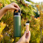 A person holds a green insulated water bottle in one hand and a small tube labeled Drinkits in the other, with leafy plants and trees in the background.