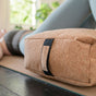 A person wearing light blue leggings lies on a yoga mat with a Yoloha Yoga Cork Bolster in the foreground. The scene is calm, illuminated by natural light with greenery in the background.