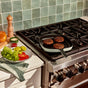 Three grilled burger patties in a grill pan on a stovetop, with a plate nearby holding lettuce, tomato, pickles, sliced onion, sauces, and a bun. Background features green tiled backsplash and a pepper grinder.