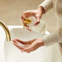 Wearing a beige sweater, a person pumps liquid soap from Grove Co.s limited edition Eves of Enchantment hand soap dispenser with a gold pump into their palm over a white sink. A gold faucet is visible, and the softly blurred background hints at festive scents in the air.