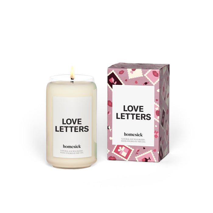 A Homesick Love Letters white candle sits beside a pink box with postage stamps, hearts, and envelopes.