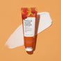 A tube of VOESH New York Velvet Luxe Vegan Hand Crème in Pumpkin Pie scent is placed on an orange background with a smear of white cream behind it. The tube features autumn leaves in red and yellow.