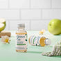 Two small bottles of Grove Co. glass cleaner concentrate sit on a light green surface, one upright and one lying down, surrounded by green apples, a flower, and a checkered cloth. White tiled wall in the background.