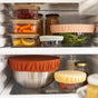 Inside a refrigerator, Esembly Babys vibrant elastic Bowl Caps cover various glass containers storing fresh produce like raspberries, carrots, lemons, and blueberries. Neatly arranged on two shelves are a mix of glass jars and covered bowls crafted from recycled plastic.