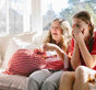 Two girls sit on a couch in sunlight, laughing with excitement. One holds a large red-and-white striped gift bag, while the other covers her mouth with her hands in surprise.