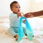 A baby in a light blue outfit reaches for Sarahs Silks Silk and FSC-Certified Wood Baby Teether, featuring colorful fabric tied to the non-toxic teether held by an adult. The softly lit background creates a calm atmosphere.