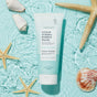 A tube of Voesh Sugar Scrub + Bubble Wash lies on a light blue surface surrounded by seashells and starfish, creating a beach-inspired scene.