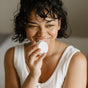 A smiling person with curly hair holds a round white container close to their mouth, wearing a sleeveless white top, sitting indoors with a soft background.