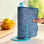 A hand places a Grove Co. Reusable “Paper” Towel, featuring blue organic cotton flannel with white wave patterns, onto a teal kitchen stand. Nearby, a wire basket of yellow lemons sits on a wooden counter, with a sink and greenery in the background.