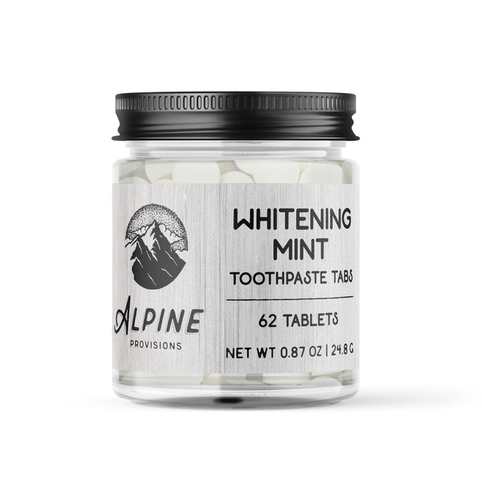A jar of Alpine Provisions Whitening Mint Toothpaste Tabs. The clear jar has a black lid, contains 62 white tablets, and the label shows a mountain logo and product details including weight: 0.87 oz (24.8 g).