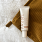 A tube of PAUME All-in-One Cuticle & Nail Cream, enriched with biotin and plant keratin, elegantly rests on a white surface and textured brown fabric. The tube is white with pink lettering, while a PAUME tag is attached to the fabric.