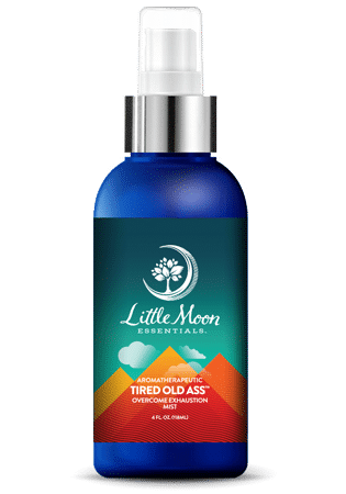 A blue spray bottle labeled Little Moon Essentials Aromatherapeutic Tired Old Ass Overcome Exhaustion Mist with a white pump and a colorful label featuring mountains and a tree logo.