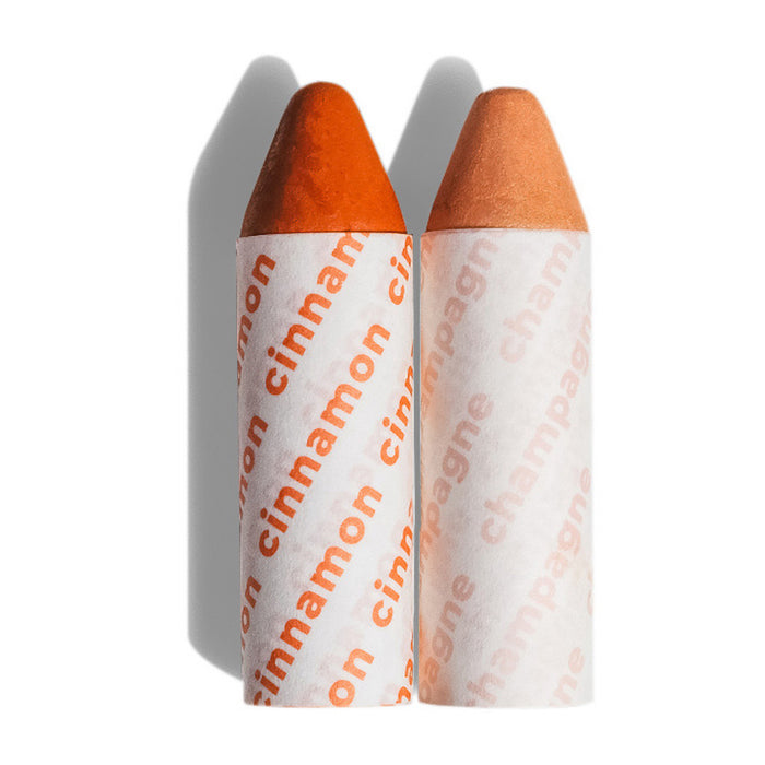 Two crayon-shaped lip balms stand upright side by side. One is labeled cinnamon in orange text and has a deep orange tip; the other is labeled champagne in peach text with a lighter, peach-colored tip.