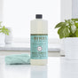 A bottle of Mrs. Meyers Clean Day Multi-Surface Concentrate in Basil scent sits on a white countertop beside a folded blue cloth and a green potted plant. The background is a bright, clean kitchen.